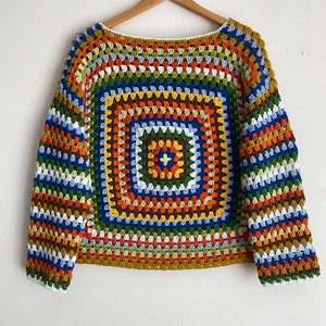 May include: A colorful crocheted sweater with a large square pattern in shades of blue, green, orange, and brown. The sweater has long sleeves and a round neckline.