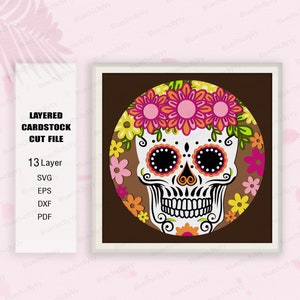 May include: A layered cardstock cut file of a sugar skull with a floral wreath. The skull is white with black eyes, a black nose, and a black mouth. The floral wreath is made up of pink, yellow, orange, and green flowers. The background is brown.