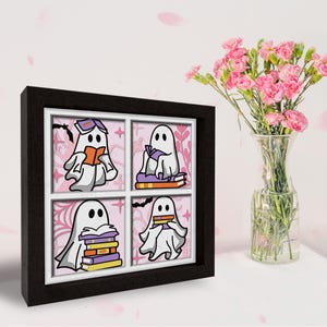 May include: A framed print with four illustrations of ghosts reading books. Each ghost is white with a purple outline and is sitting on or holding a stack of books. The background is pink with a floral pattern.