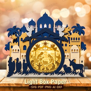 May include: A multi-layered paper light box featuring a nativity scene inside a circular cutout. The design includes dark blue and tan architectural elements, palm trees, and two glittery silver angels. The text reads Light Box Paper.