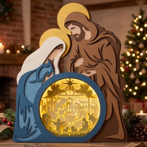 May include: A multi-layered wooden nativity scene featuring stylized figures of Mary in blue and Joseph in brown, with gold halos. A central circular cutout displays an illuminated silhouette of the manger, angels, and animals against a warm light.