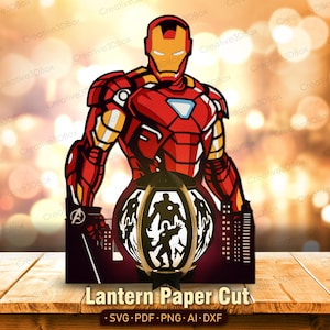 Può includere: Un design in carta tagliata a strati che raffigura Iron Man nella sua armatura rossa e oro. Sotto il personaggio si trova una struttura a lanterna circolare con sagome dell'eroe, su uno sfondo di skyline cittadino con il logo Avengers.