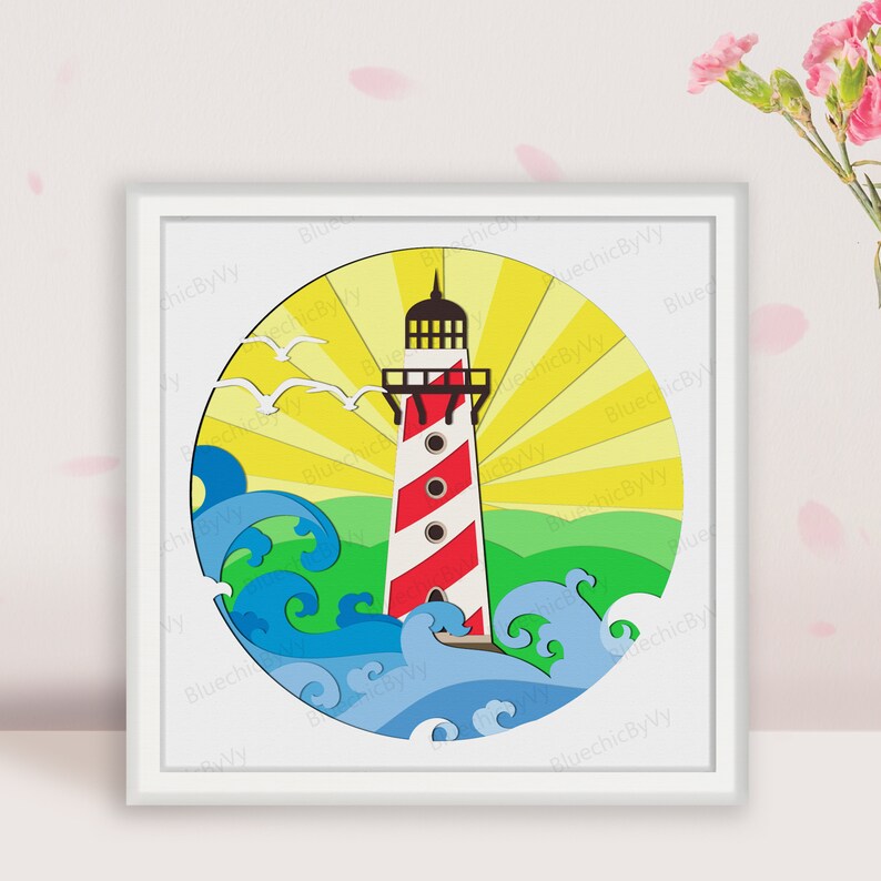 May include: A framed, layered paper art piece featuring a lighthouse with red and white stripes, set against a yellow sunburst. Blue waves and green land are at the base, with white seagulls in the sky. The artwork is in a white frame.