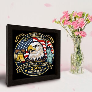 May include: A framed artwork commemorating the 250th anniversary of the United States of America. The design includes an eagle, the American flag, the Liberty Bell, and the Statue of Liberty. The frame is dark brown, and a vase of pink flowers is next to it.