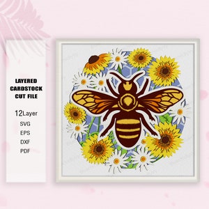 Può includere: Un file di taglio in cartoncino a strati con un'ape circondata da girasoli e margherite. L'ape è marrone e gialla con strisce nere. I fiori sono gialli e bianchi con foglie verdi.