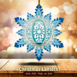 May include: A blue and white paper snowflake lantern with a geometric design. The lantern is hanging from a string and is in front of a blurred background of golden lights.