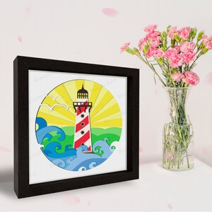 May include: A framed artwork featuring a lighthouse with red and white stripes, set against a yellow sunburst and blue waves. The artwork is in a black frame. The image also includes a vase of pink flowers.