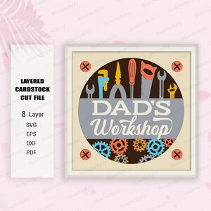 May include: A layered cardstock cut file design for a workshop sign. The sign is a circle with the text "DAD'S Workshop" in a gray circle with a brown background. The circle is surrounded by gears and tools in various colors, including red, blue, yellow, and green. The sign is framed in a white frame with an X in each corner.