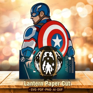Op de afbeelding: Een meerlaags papierontwerp met Captain America in zijn blauw, wit en rood kostuum met zijn ronde schild. Daaronder toont een donkere lantaarnstructuur silhouetten van superhelden tegen een stadssilhouet, met de tekst Lantern Paper Cut.