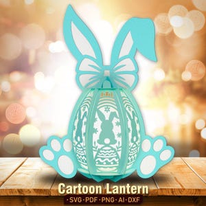 Pu&ograve; includere: Una lanterna di Pasqua a forma di coniglio blu chiaro con un fiocco bianco e un design a uovo bianco. La lanterna ha la forma di un uovo e ha un viso di coniglio bianco sul davanti. La lanterna &egrave; appoggiata su una superficie di legno.