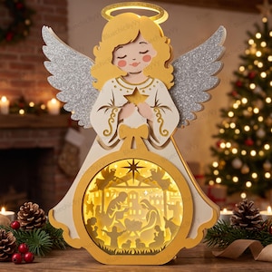 May include: A layered paper angel decoration featuring a yellow halo, glittery silver wings, and a central circular cutout displaying a backlit nativity scene with figures of people and animals in warm yellow light.