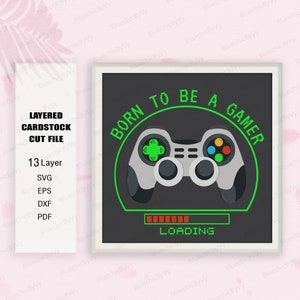 May include: A black and green layered cardstock cut file design with the text "Born to be a gamer" in a circle around a gray video game controller. The controller has green buttons and a green progress bar below it that reads "Loading".