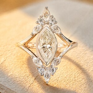Op de afbeelding: Een marquise-geslepen diamanten ring met een uniek ontwerp. De ring heeft een grote, centrale marquise-diamant, omringd door kleinere, ronde diamanten. De zetting is een delicaat, geometrisch ontwerp in een licht metaal.