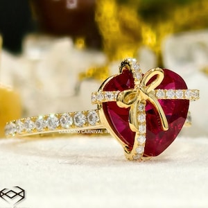 May include: A gold ring featuring a red heart-shaped gemstone set in the centre. The heart is wrapped with a gold ribbon and bow, and the band is set with small diamonds. "DIAMOND CARNIVAL IN" is printed on the band.