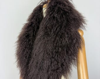 May include: A dark brown, long-haired, shaggy fur collar draped over a wooden mannequin. The accessory features a thick, textured pile with a natural, uneven appearance, creating a voluminous and layered look around the neck area.