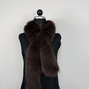 Puede incluir: Bufanda de piel de color marrón oscuro exhibida en un maniquí negro. La bufanda está drapeada alrededor del cuello y cuelga en dos secciones largas. La piel parece suave y lujosa, con un color rico y oscuro.