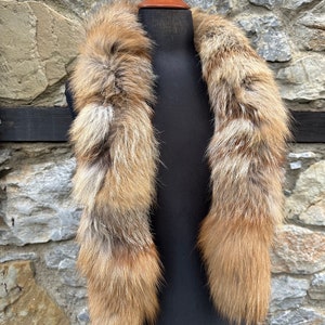 May include: A long, brown and white fur collar with a fluffy texture. The collar is displayed on a black mannequin against a stone wall background.