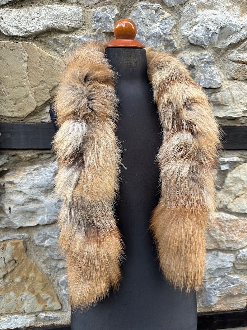 May include: A long, brown and white fur collar with a fluffy texture. The collar is displayed on a black mannequin against a stone wall background.