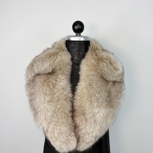 Op de afbeelding: Een luxe, lichtbruine bontkraag met een zachte, getextureerde uitstraling. De kraag wordt getoond op een zwarte mannequin, die de vorm en het volume laat zien. De vacht heeft een natuurlijke, multi-tone kleurvariatie, wat diepte aan het ontwerp toevoegt.