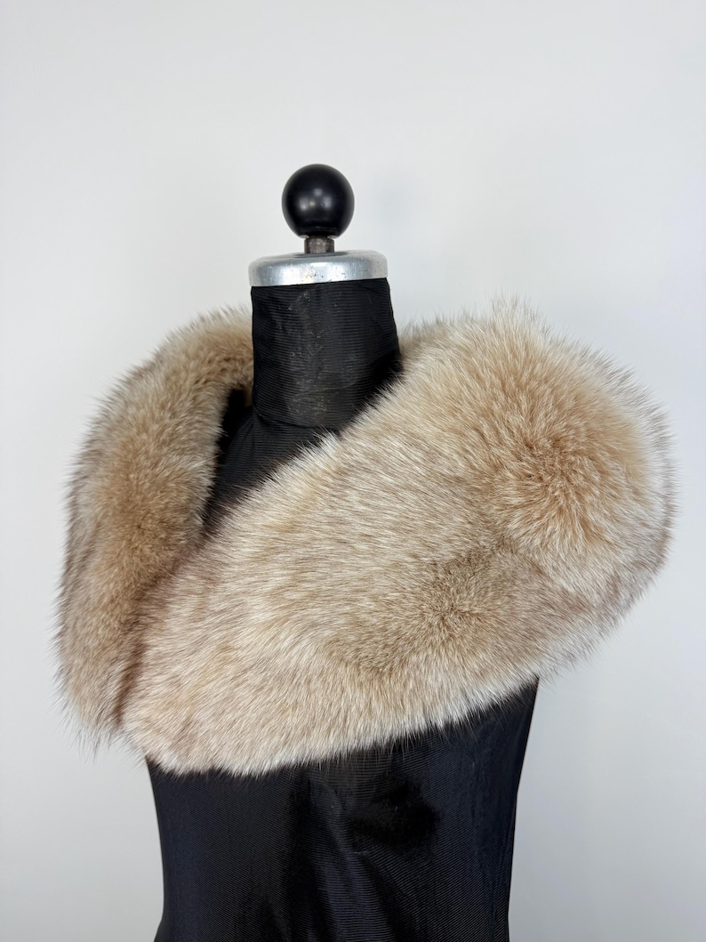Op de afbeelding: Een luxe, lichtbruine bontkraag, tentoongesteld op een zwarte mannequin. De vacht heeft een zachte, donzige textuur met lichtbruine en grijze tinten. De kraag is ontworpen om om de nek te worden gedragen.
