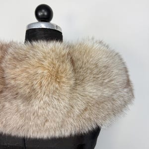 Op de afbeelding: Een luxe, lichtbruine bontkraag, gepresenteerd op een zwarte mannequin. De vacht heeft een zachte, donzige textuur en een natuurlijk kleurverloop, vari&euml;rend van lichtbeige tot donkerdere bruine punten. De kraag is ontworpen om om de nek te worden gedragen.