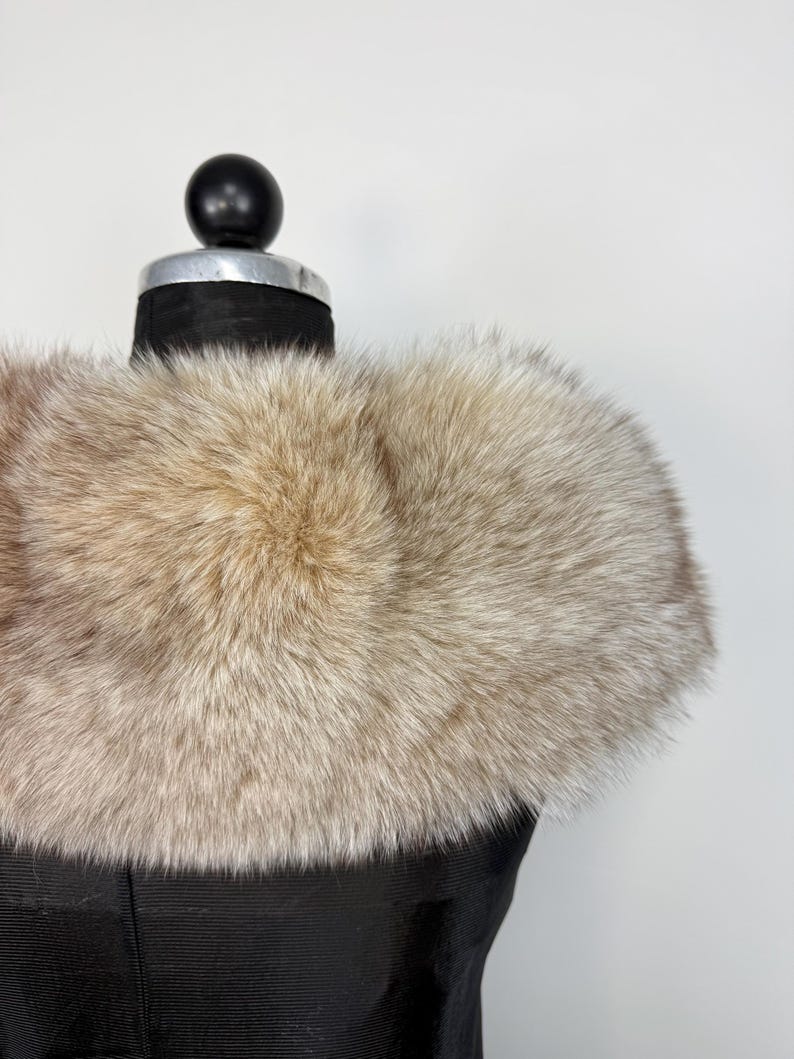 Op de afbeelding: Een luxe, lichtbruine bontkraag, gepresenteerd op een zwarte mannequin. De vacht heeft een zachte, donzige textuur en een natuurlijk kleurverloop, vari&euml;rend van lichtbeige tot donkerdere bruine punten. De kraag is ontworpen om om de nek te worden gedragen.