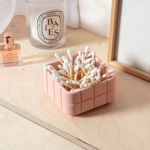 Peut inclure: Un contenant carré rose avec un motif en grille contient une grande quantité de cotons-tiges. Le contenant est posé sur une surface en bois clair.