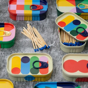 May include: Colorful, patterned tins with pull-top lids, resembling sardine cans, are arranged on a gray surface. Some tins are open, revealing wooden matchsticks with blue tips. The tins feature geometric designs in various colors, including red, yellow, and blue.
