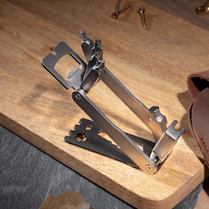 Könnte beinhalten: Ein silbernes Multitool mit einem Flaschenöffner, einem Schraubendreher und anderen Werkzeugen. Das Werkzeug ist zusammengeklappt und liegt auf einer Holzoberfläche.