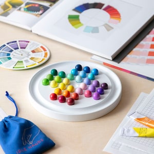 May include: A white circular game board with a rainbow of coloured marbles arranged in a circular pattern. The board is on a light wood surface.