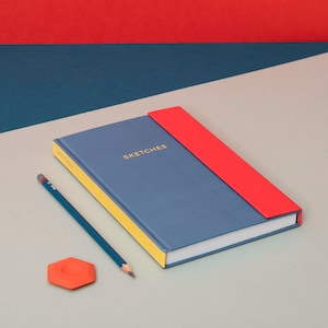 Pode incluir: Um caderno de esboços azul com uma lombada amarela e um marcador vermelho. O caderno de esboços tem a palavra "SKETCHES" impressa na capa. Um lápis e uma borracha vermelha estão sobre uma superfície cinza claro.
