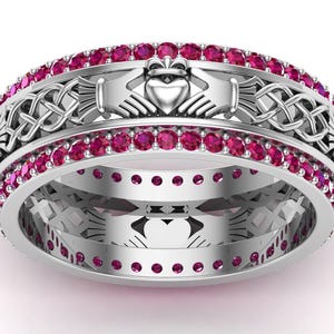 Peut inclure: Une bague en argent avec un motif de Claddagh, représentant un cœur tenu par deux mains et des nœuds celtiques. La bague est ornée d'une rangée de petites pierres roses rondes. Le motif de la bague est répété sur la bande intérieure.