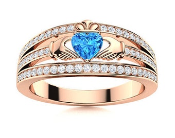May include: A rose gold ring with a blue heart-shaped gemstone surrounded by a halo of smaller diamonds. The ring features a Celtic knot design with two hands holding the gemstone.