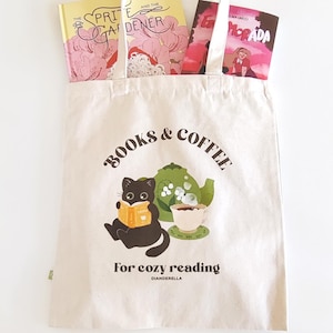 Puede incluir: Una bolsa de lona blanca con el texto "BOOKS & COFFEE for cozy reading" y un gato negro leyendo un libro. La bolsa tiene una tetera y una taza verdes con vapor saliendo de ella.