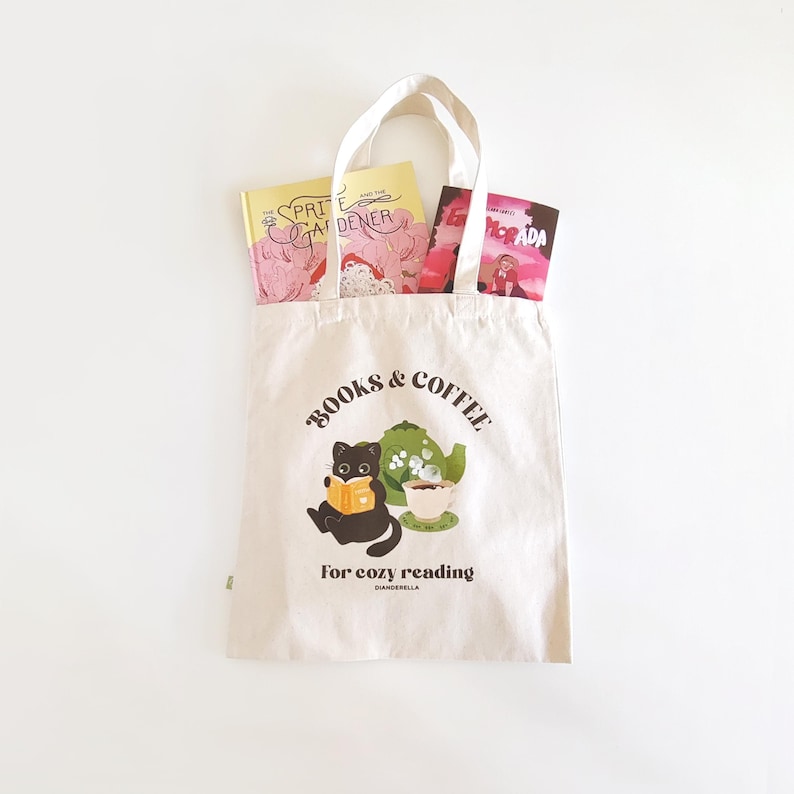 Puede incluir: Una bolsa de lona blanca con el texto "BOOKS & COFFEE for cozy reading" y un gato negro leyendo un libro. La bolsa tiene una tetera y una taza verdes con vapor saliendo de ella.
