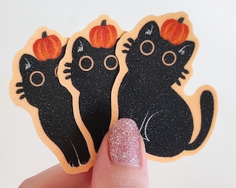 May include: Three black cat stickers with orange pumpkins on their heads. The stickers are on a peach-colored background.