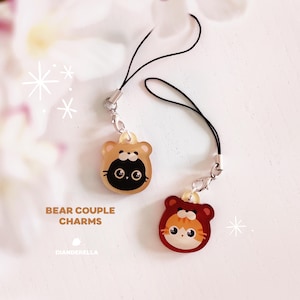 Puede incluir: Dos encantos de pareja de osos lindos con caras de gato negro y naranja. Los encantos están unidos a correas de teléfono negras con cierres plateados. Los encantos están etiquetados como "BEAR COUPLE CHARMS" y "DIANDERBELLA".