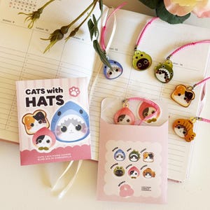 Peut inclure: Ensemble de breloques de téléphone "Chats avec chapeaux" et emballage de boîte aveugle. Les breloques présentent des illustrations de chats avec des chapeaux, dont un requin, du pain grillé et une pêche. L'emballage est rose avec du texte et des illustrations blancs.