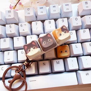 Pode incluir: Um porta-chaves com quatro teclas de teclado quadradas dispostas em fila, cada uma decorada com uma cara de gato de desenho animado. Os gatos incluem um gato preto e branco, um gato preto, um gato tricolor e um gato laranja. Anel de metal castanho.