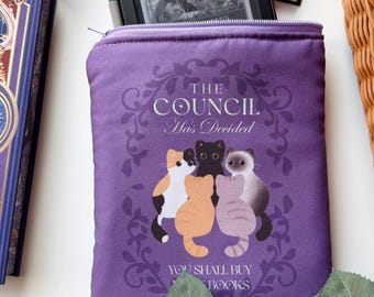 Pode incluir: Uma bolsa de tecido roxa com fecho de correr, apresentando um gráfico de cinco gatos dispostos em círculo. O texto diz THE COUNCIL Has Decided e YOU SHALL BUY MORE BOOKS. A bolsa é retangular com uma borda floral roxa escura.