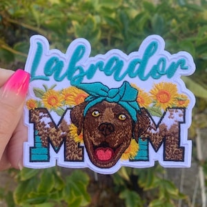 May include: White embroidered patch with the words "Labrador Mom" and a brown Labrador Retriever wearing a turquoise bandana. The patch is surrounded by yellow sunflowers.