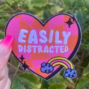 May include: Heart-shaped patch with the words "EASILY DISTRACTED" in white embroidery. The patch has a pink and orange checkered background, a rainbow, and two black starbursts. The patch is a fun accessory.