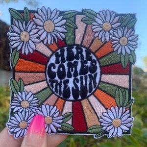 May include: Embroidered patch with a retro sunburst design in shades of orange, red, and green. The center of the patch features a white circle with the text "Here Comes The Sun" in black. The patch is surrounded by white daisy flowers.