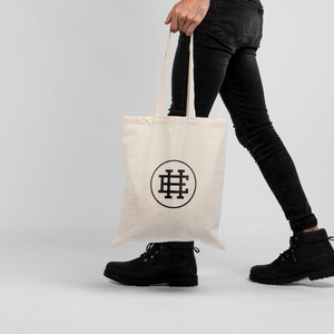 May include: A person carrying a white tote bag with a black logo of a stylized "H" in a circle. The bag has a long strap and is made of canvas.