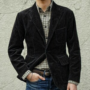 May include: A black corduroy blazer with a notched lapel and two front pockets. The blazer is worn over a plaid button-down shirt and blue jeans. The person is wearing a brown belt and a silver bracelet.