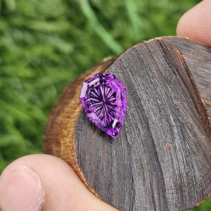 May include: A vibrant purple gemstone with a shield-shaped cut and intricate radial faceting that creates a star-like pattern. The stone is positioned on a circular, dark brown wooden slice with visible grain and texture, showcasing its deep color.