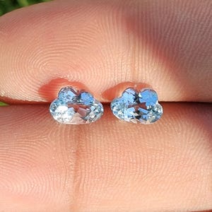 May include: Two small, faceted, light blue gemstones shaped like clouds. Each stone features a clear, translucent appearance with multiple cut surfaces that reflect light, creating a sparkling effect across the top of the cloud-shaped design.