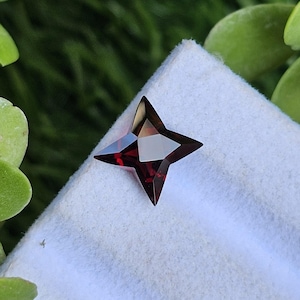 May include: A deep red, faceted gemstone cut into a four-pointed star shape. The stone features sharp, geometric edges and a polished surface that reflects light, creating distinct dark and light facets across its dark crimson, translucent body.