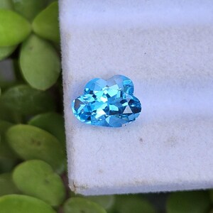 May include: A bright blue, faceted gemstone cut into a distinct cloud shape. The stone features multiple triangular and trapezoidal facets that reflect light, creating a sparkling appearance. The gemstone is positioned on a white surface.