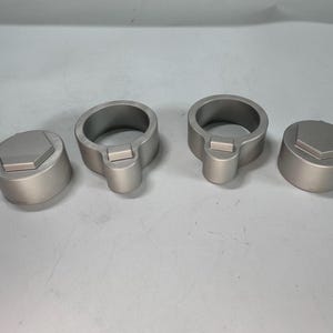 May include: Four silver metal wheel lug nut covers. Two of the covers are round with a hexagonal opening in the center. The other two covers are round with a hexagonal opening in the center and a small, rectangular opening on the side.